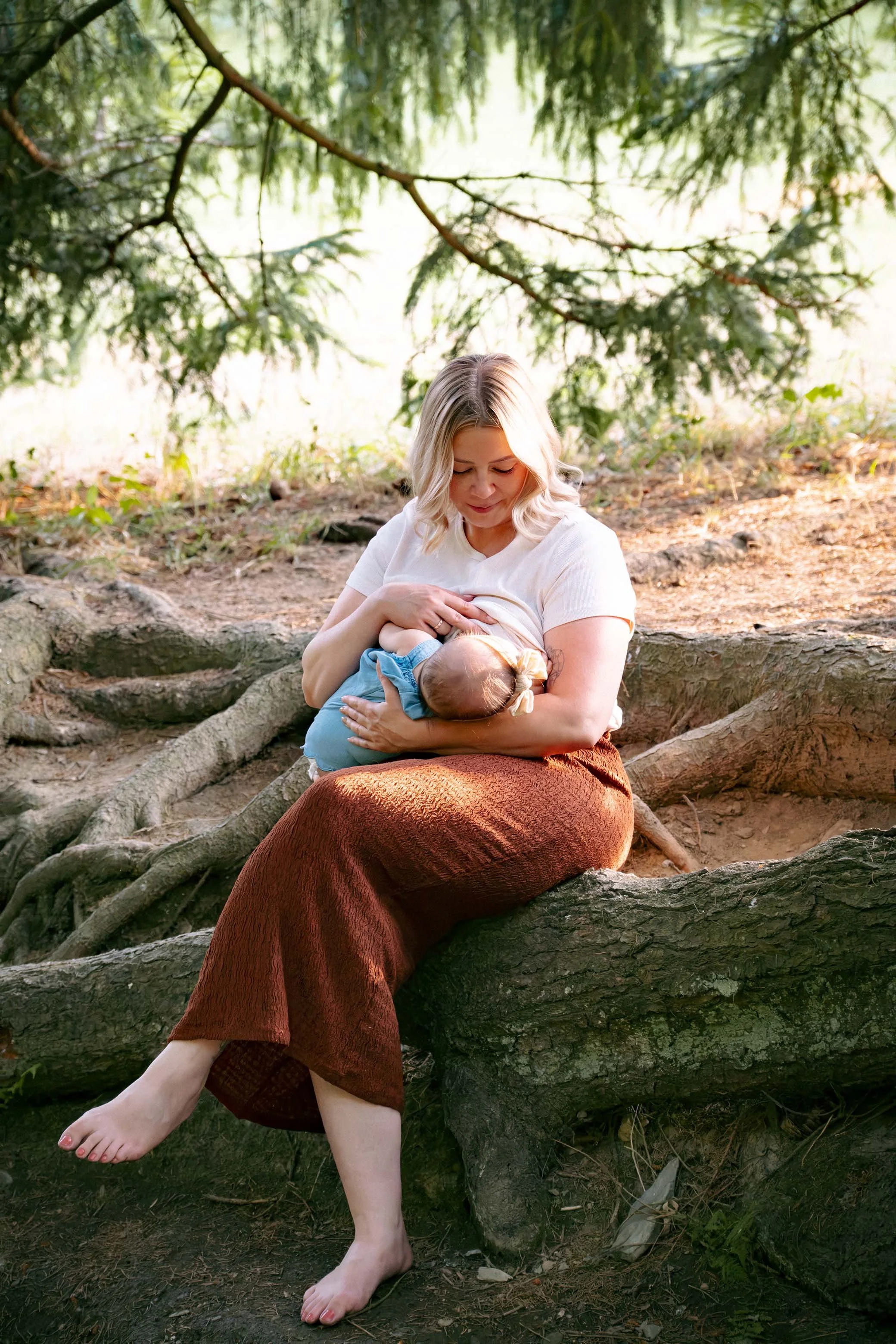 Eine Frau sitzt auf einem Baumstamm im Wald, hält ein Baby beim Stillen und lächelt sanft, während sie die Natur um sich herum genießt. Entstanden bei einem Familien Shooting in Schmitten im Taunus.