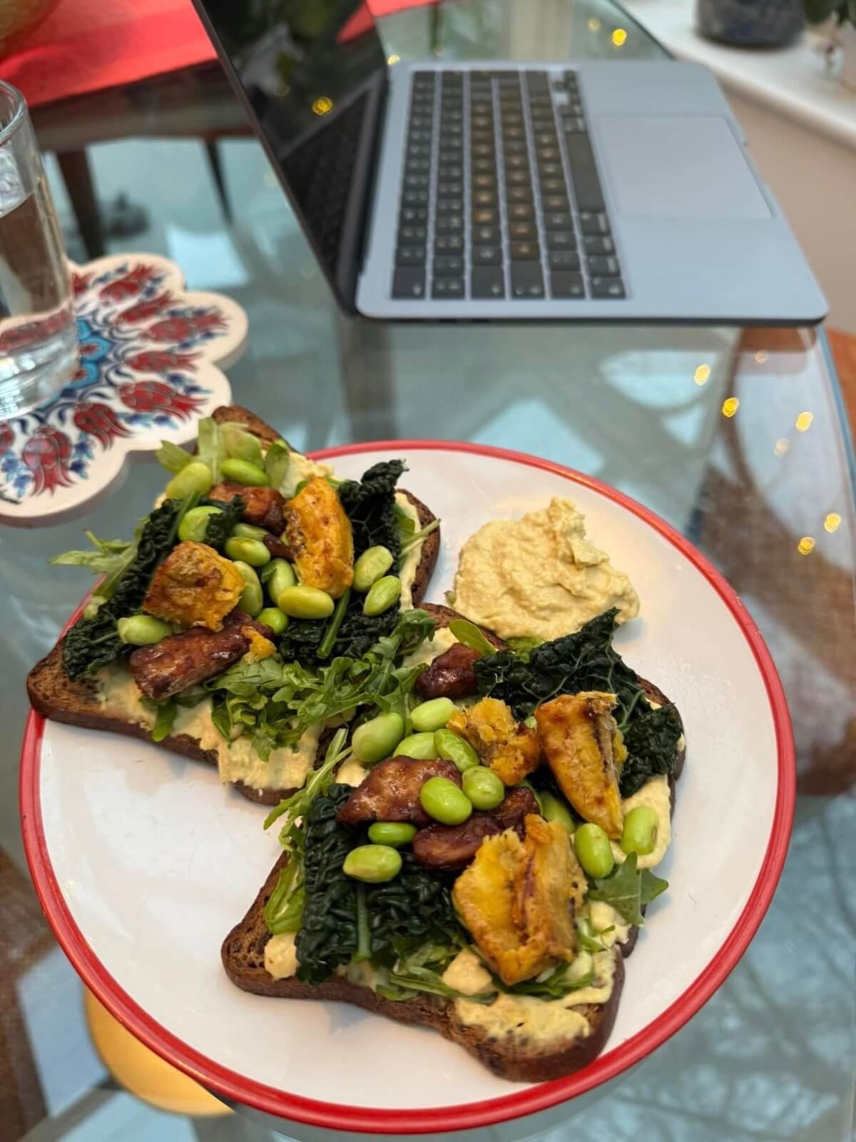 loaded toast with hummus and edamame beans next to a laptop on a plate