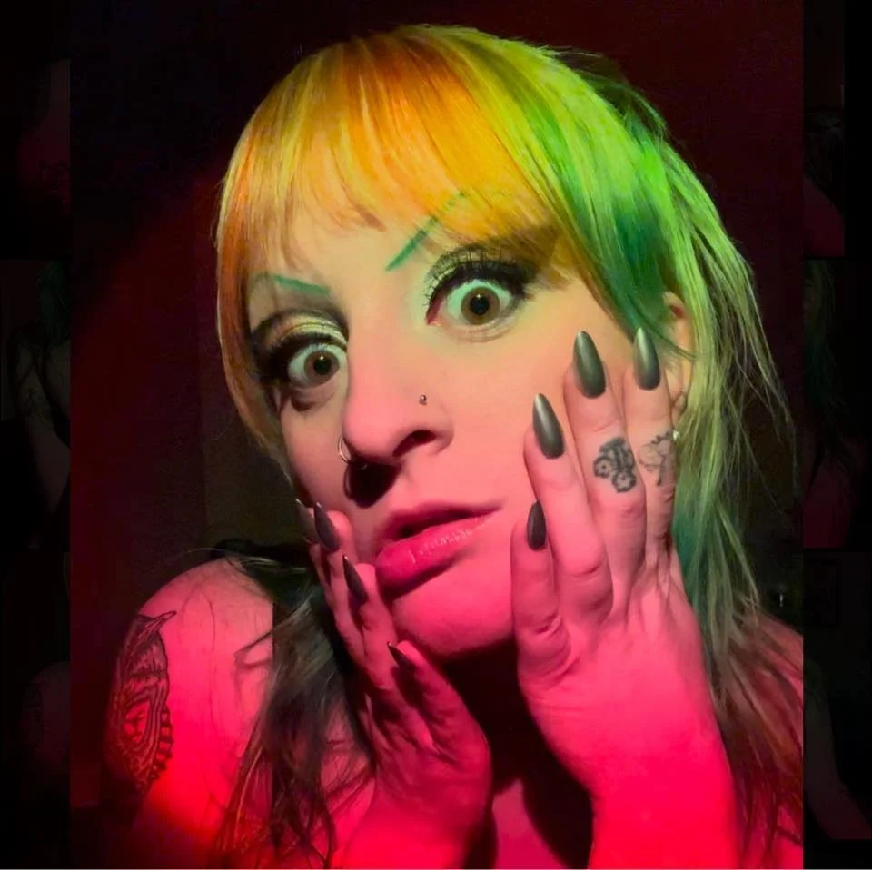 A woman with multi-colored hair, green and orange, poses with her hands on her face, showing long dark nails, tattoos, and facial piercings, with an expressive facial expression.