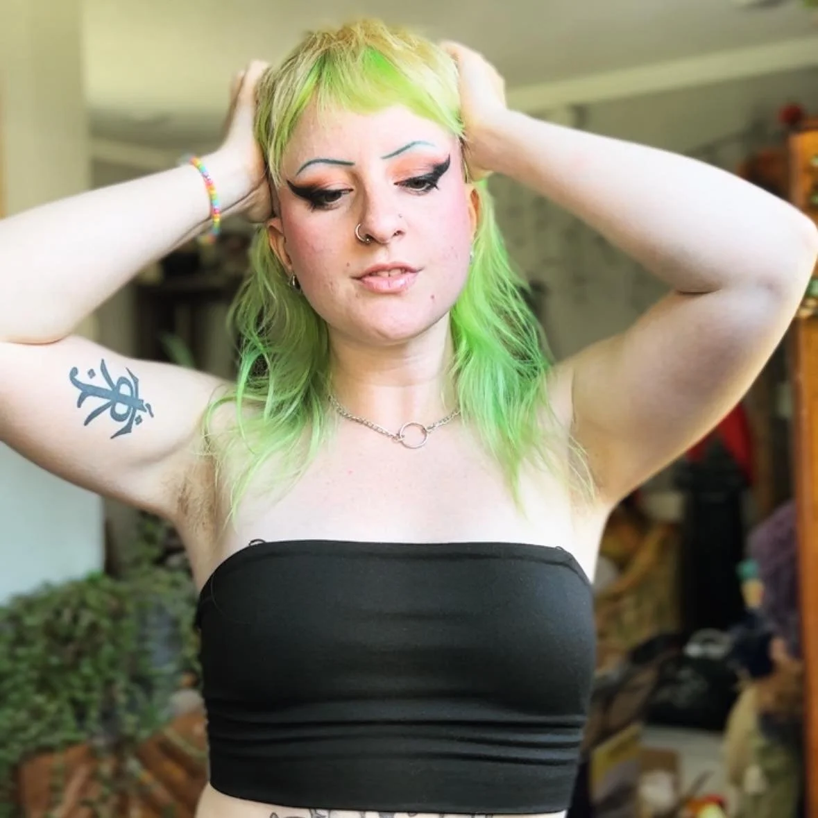 A young woman with neon green hair, wearing a black strapless top, standing indoors with her hands behind her head, showcasing makeup, tattoos, and jewelry.