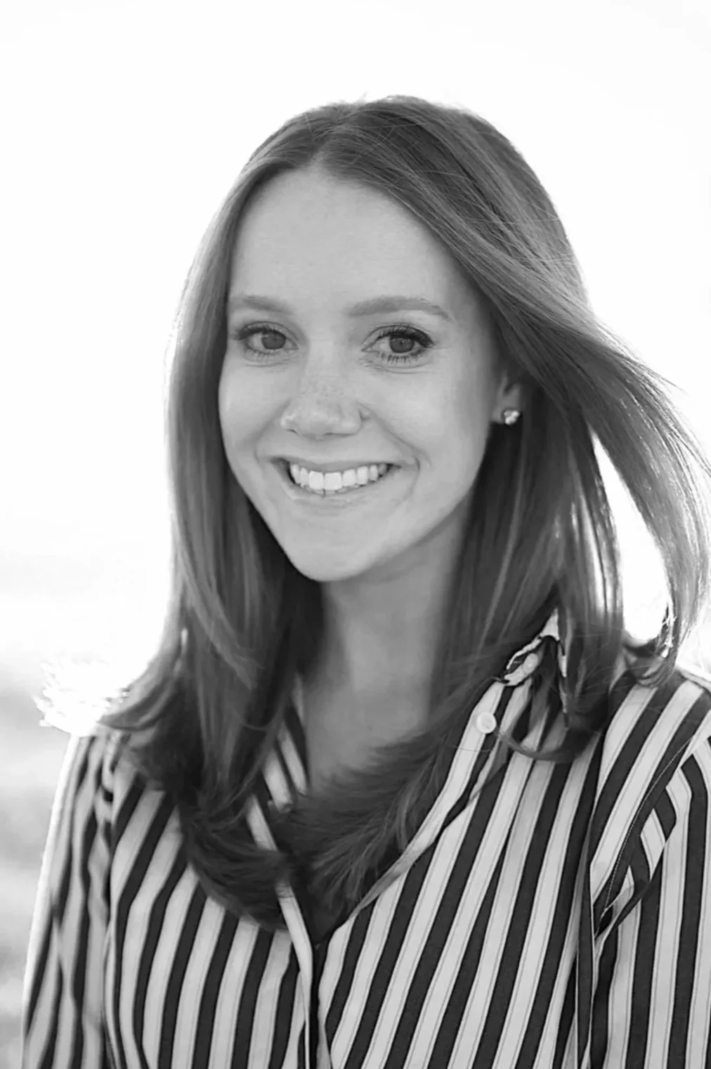 Black and white portrait of a smiling young woman with long, wavy hair, wearing a dark button-up shirt and a delicate necklace, standing indoors near a large window and bookshelf.
