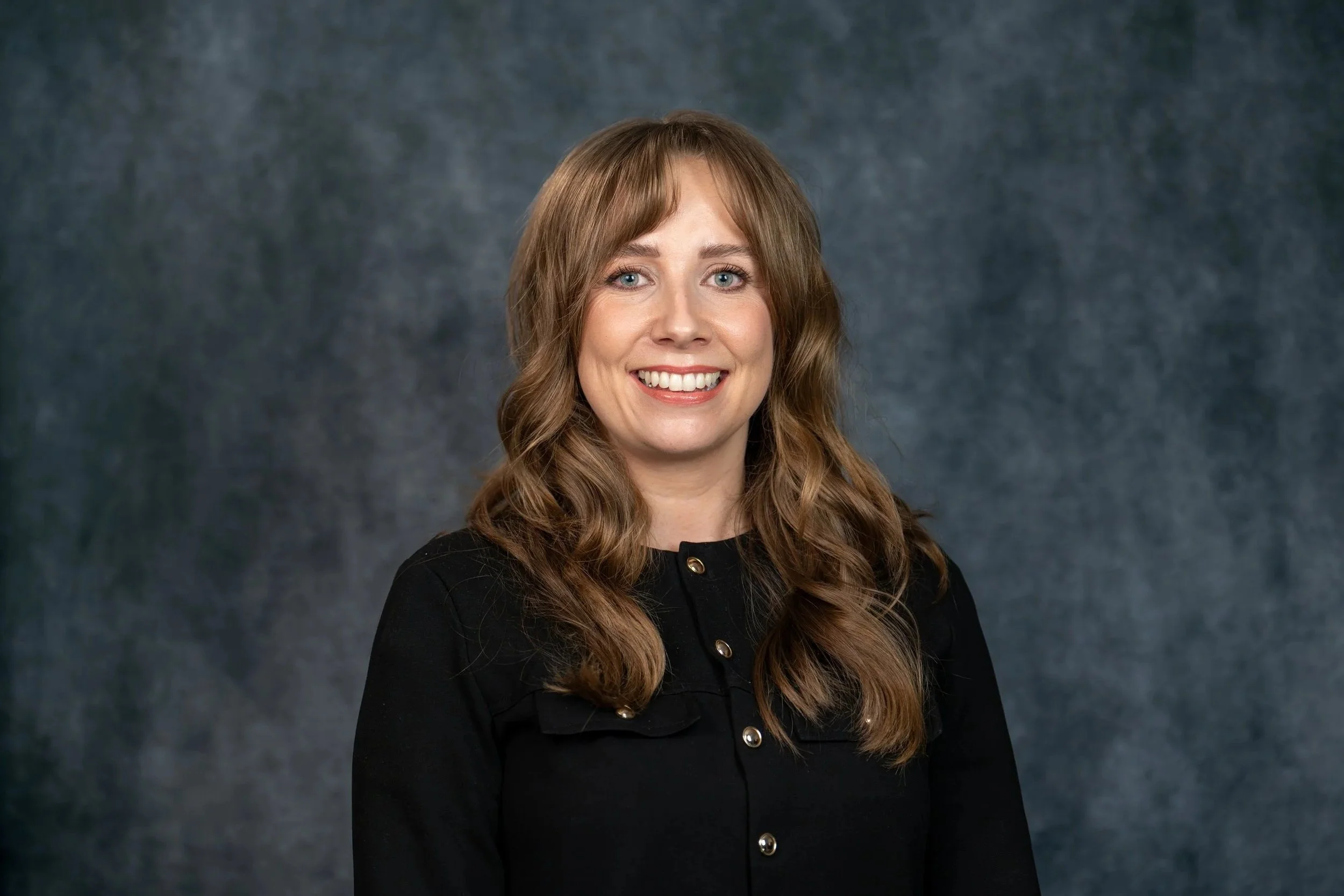 A young woman with long, wavy brown hair and light skin, smiling at the camera, wearing a black blouse and a thin gold necklace, against a gray background.