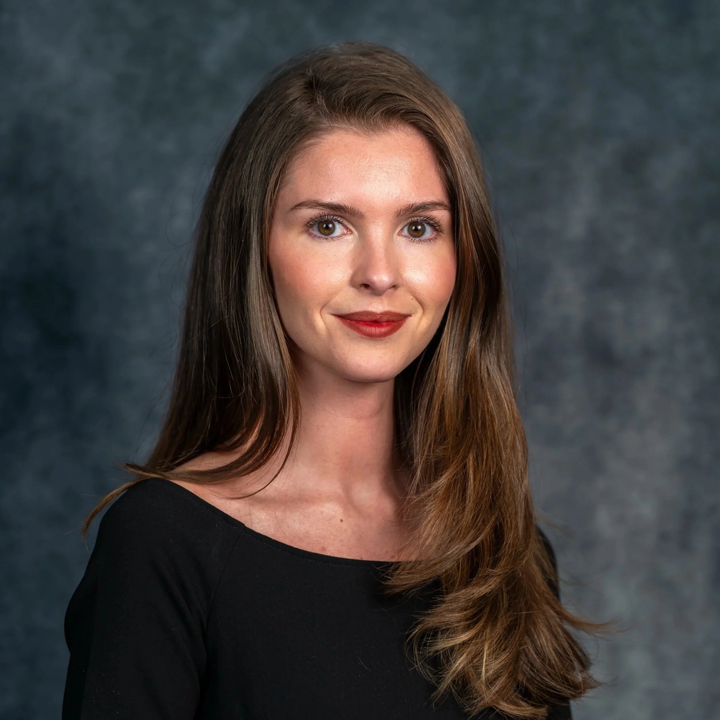 A young woman with long brown hair, smiling, wearing a black top and a delicate necklace against a gray background.
