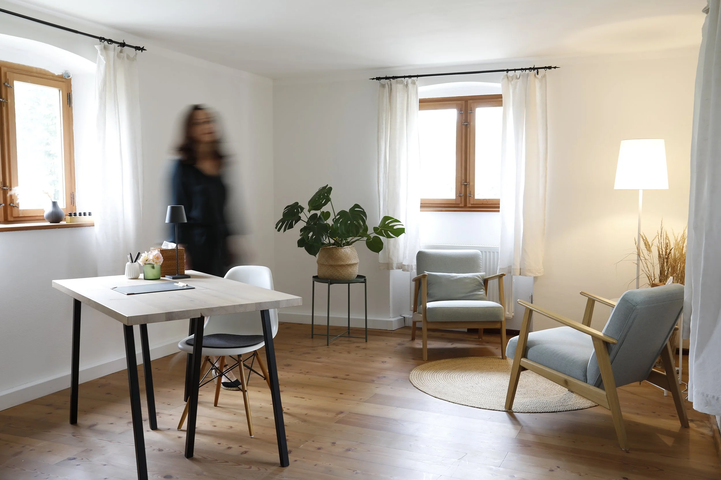 Helles Wohnzimmer mit Holzfußboden, zwei Fenster mit weißen Vorhängen, einen Tisch mit Stuhl, einen Sessel, einen Beistelltisch mit Topfpflanze und einen Stehlampe