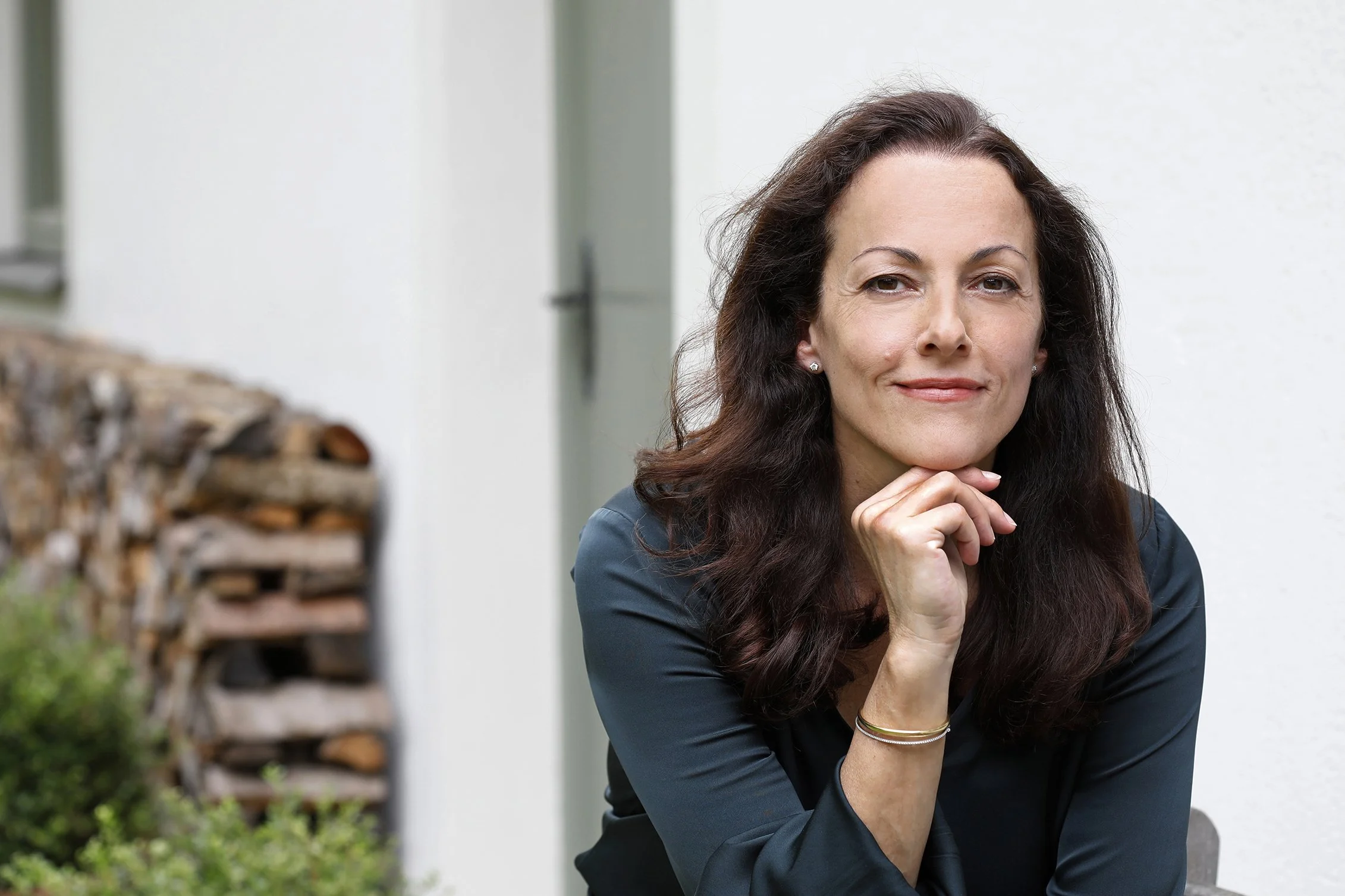 Nahaufnahme einer Frau mit dunklen, lockigen Haaren, die lächelnd nach vorne schaut, die Hand an das Kinn gestützt, im Hintergrund eine weiße Wand und Holzscheite.