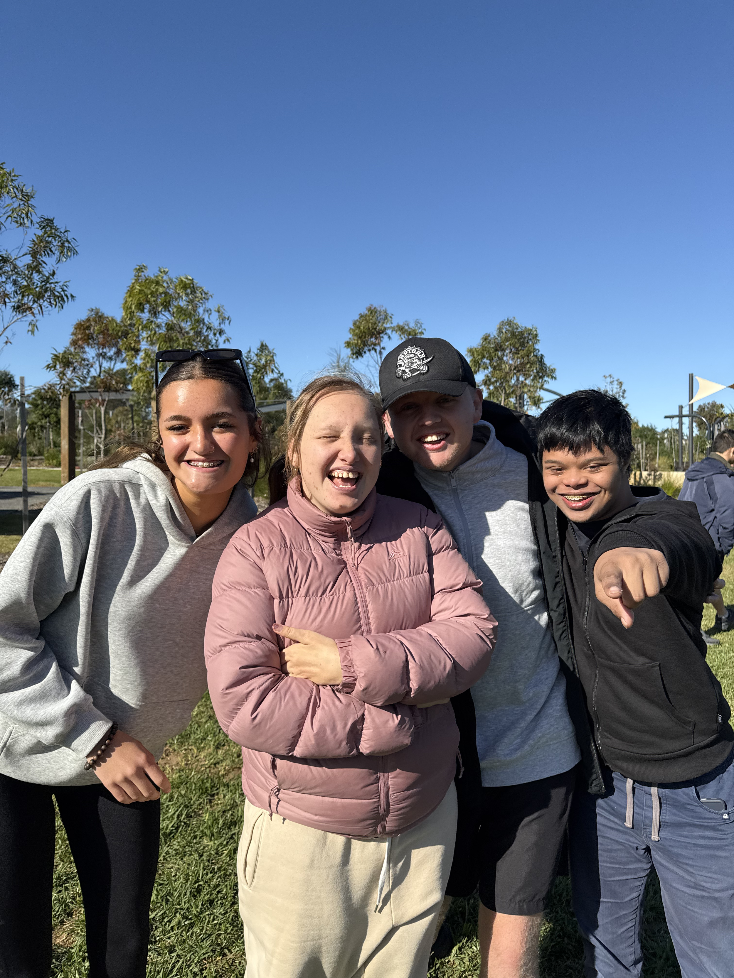 Four young people standing close together outdoors on a sunny day, smiling and posing for the camera.