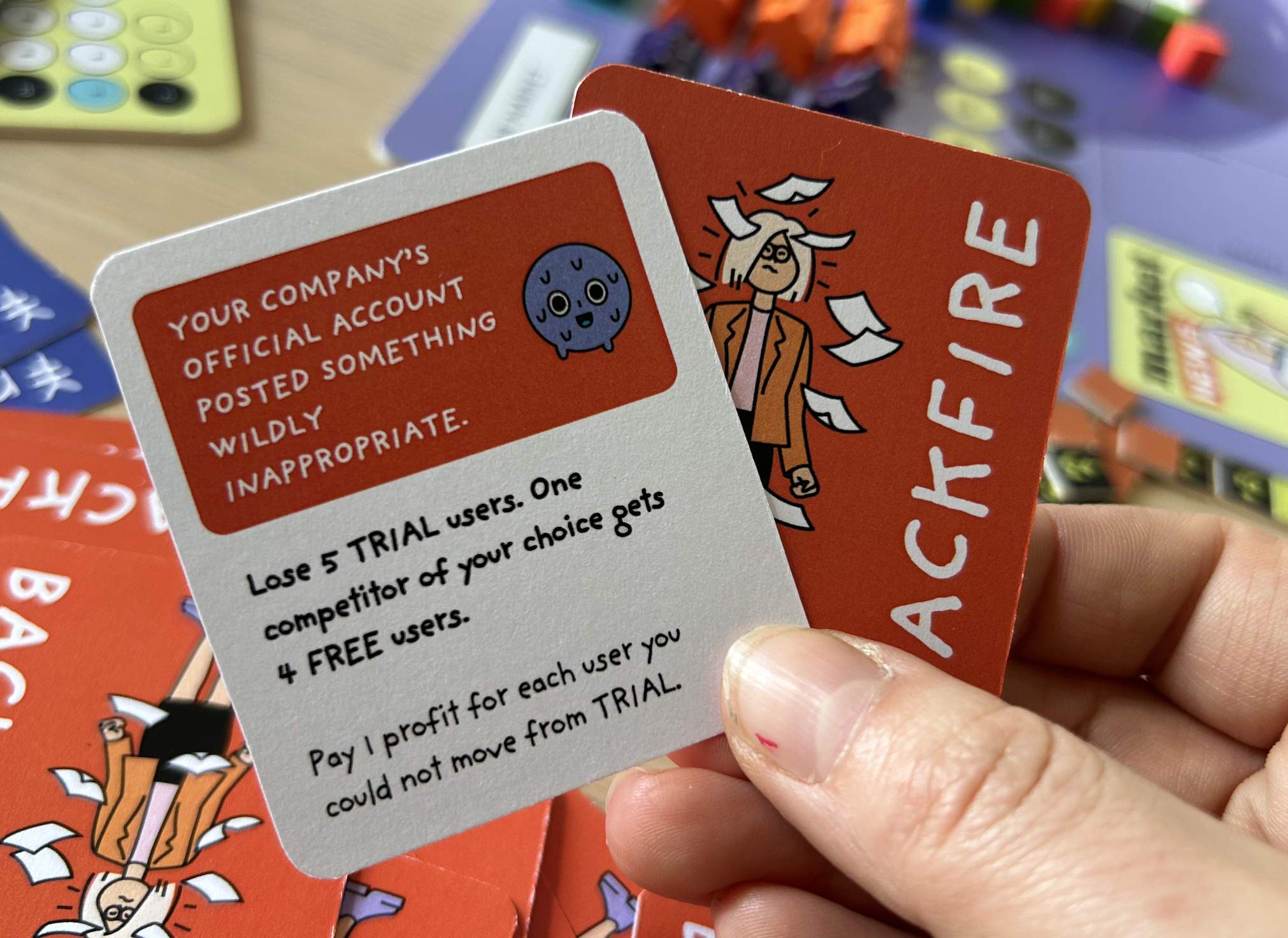 A hand holding two cards from a game called 'Backfire,' featuring comic-style illustrations and humorous instructions for players.