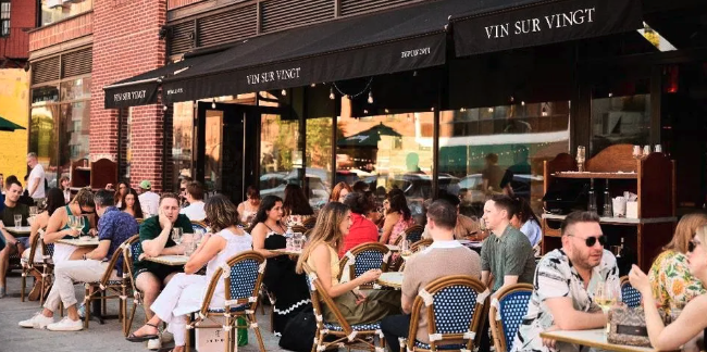People dining outside at a restaurant with a black awning that reads 'Vin Sur Vingt'