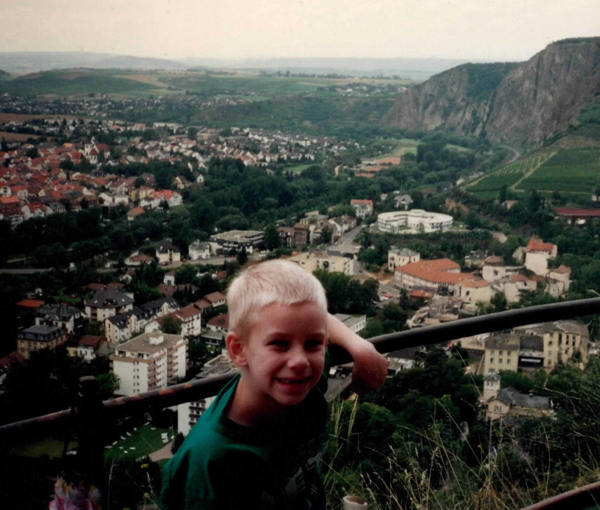 A young boy smiling and giving a thumbs-up gesture, with a cityscape and green hills in the background.