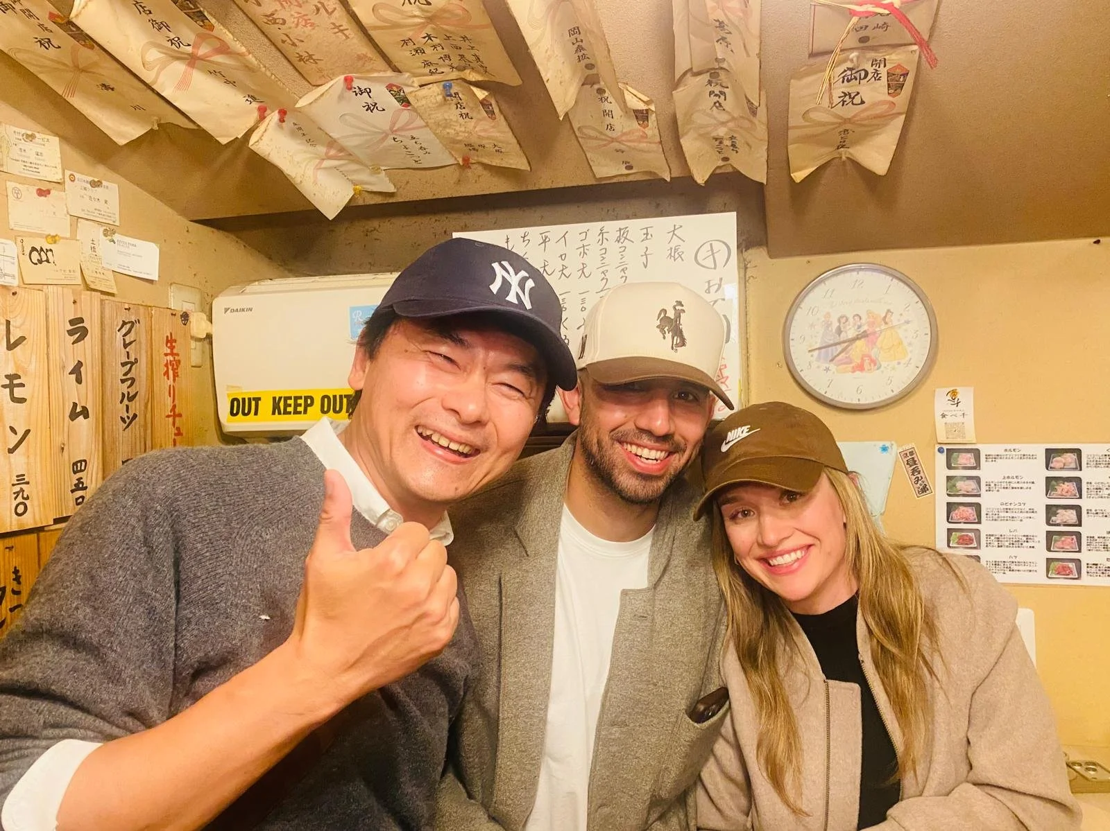 Three people smiling and posing for a photo inside a Japanese restaurant, with menus and decorations on the wall behind them.