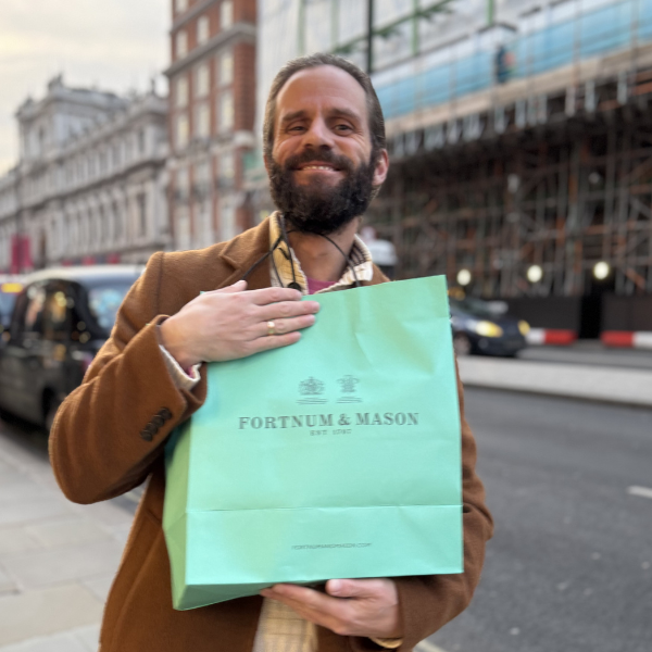 Smiling man with a beard holding a green shopping bag from Fortnum & Mason on a city street.