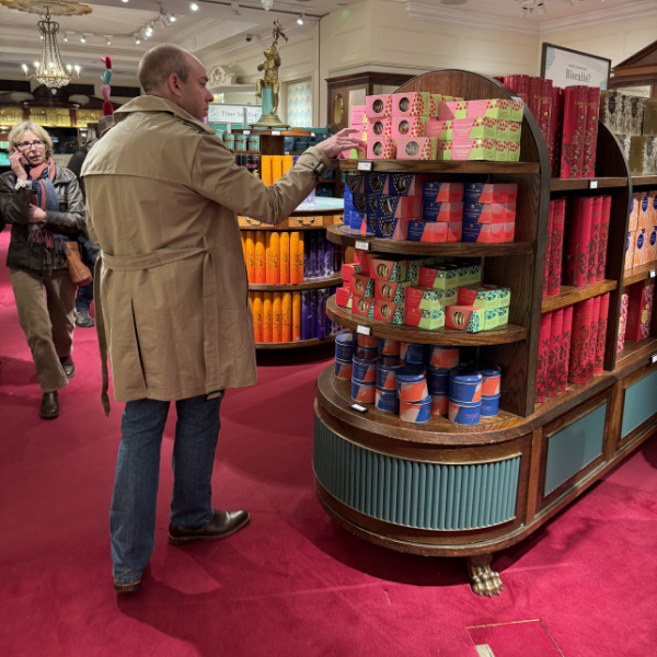 A man in a khaki trench coat shopping for gift boxes on a wooden display shelf in a store decorated with colorful packaging and ornaments. Another person is in the background.