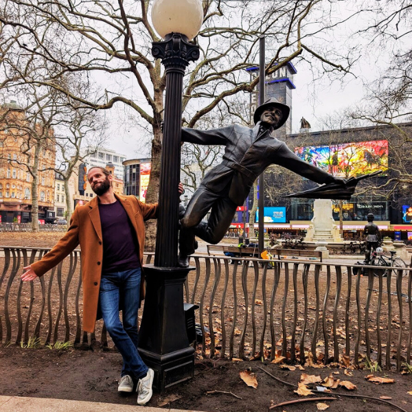 A man in a brown coat and jeans standing next to a lamp post sculpture in a park. The sculpture depicts a person reaching out with one arm, holding a book, and the other arm stretched downward. Trees without leaves and city buildings are visible in t