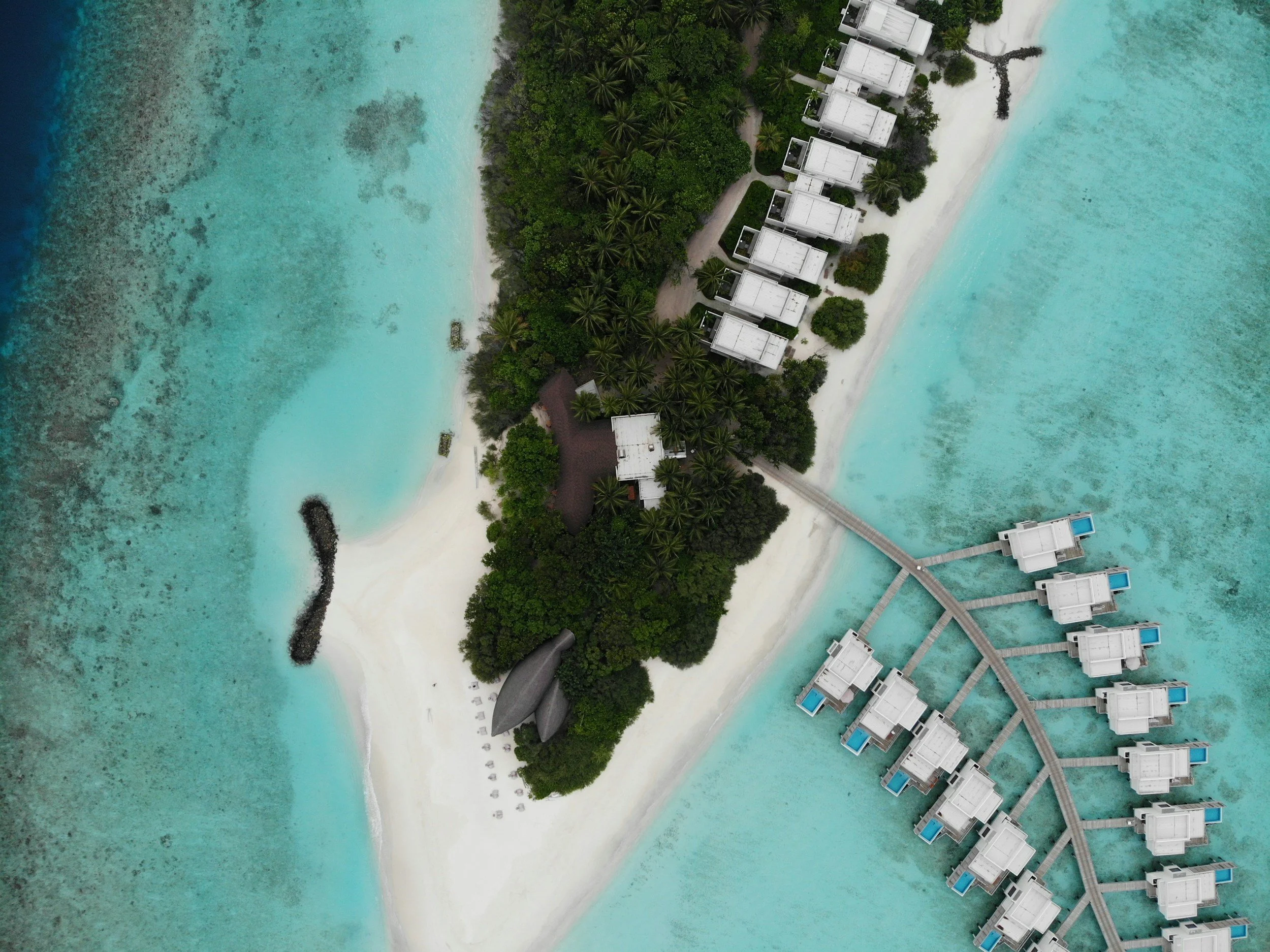 Aerial view of overwater bungalows with private pools, white sandy beach, lush greenery, and turquoise ocean water at a tropical resort.