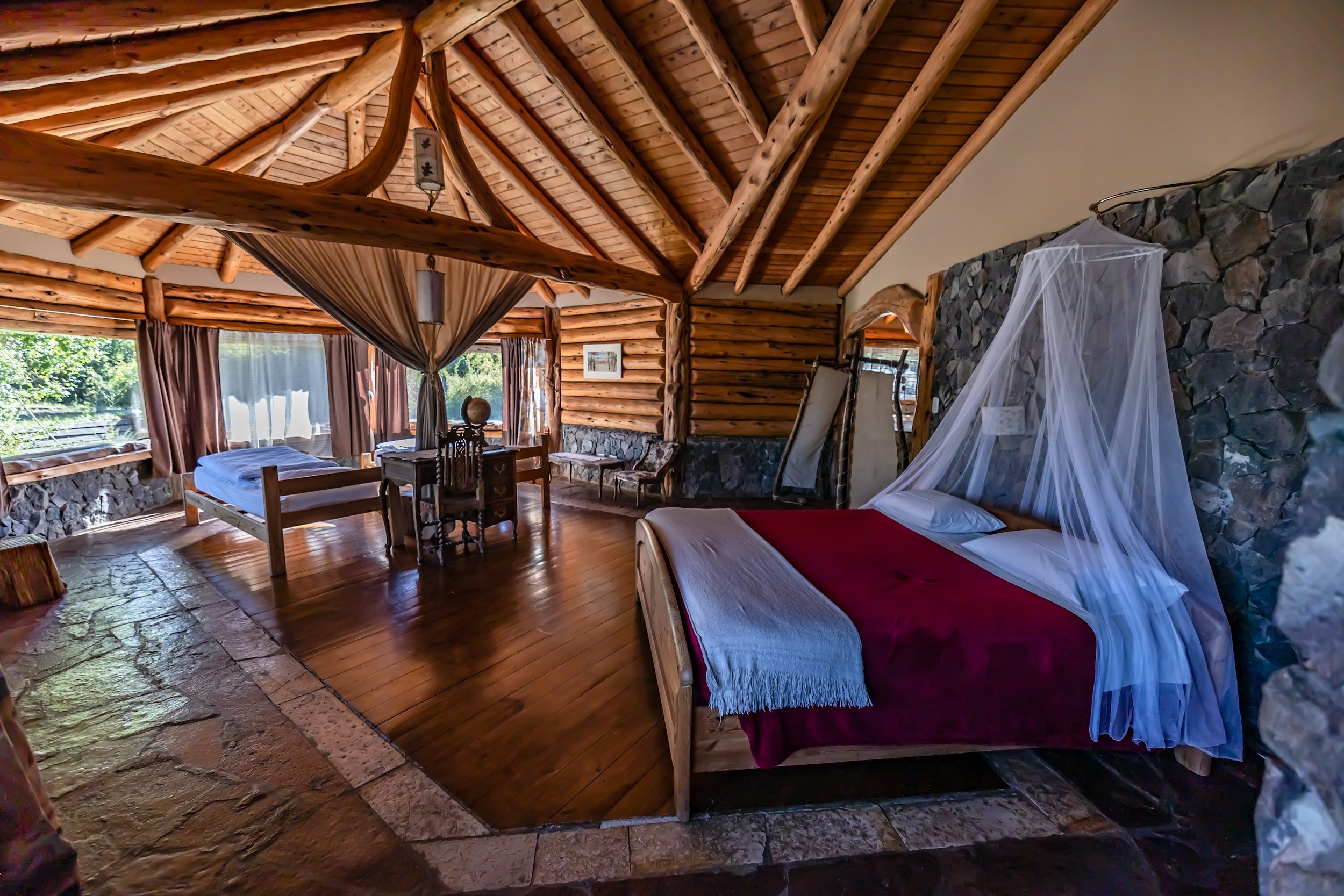 Interior of a rustic bedroom with wooden log walls and ceiling, a bed with a white mosquito net, and large windows with curtains showing greenery outside.