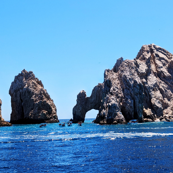 Large ocean waves with boats near rocky sea stacks with natural arch under a clear blue sky.