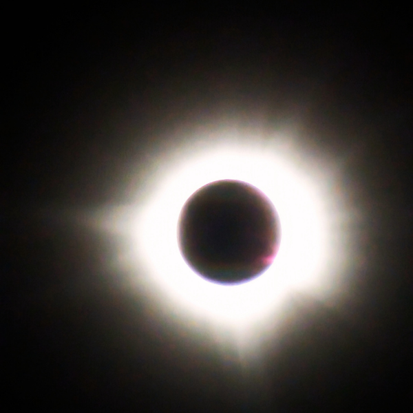 A total solar eclipse hangs in a black sky — the corona blazing around a perfect dark disc, captured at totality.