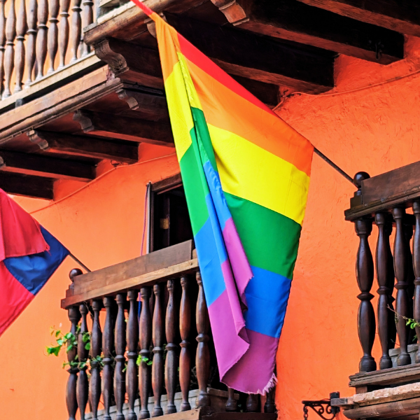 Rainbow pride flag hanging on a wooden balcony railing in front of an orange building, with another flag partially visible on the left.