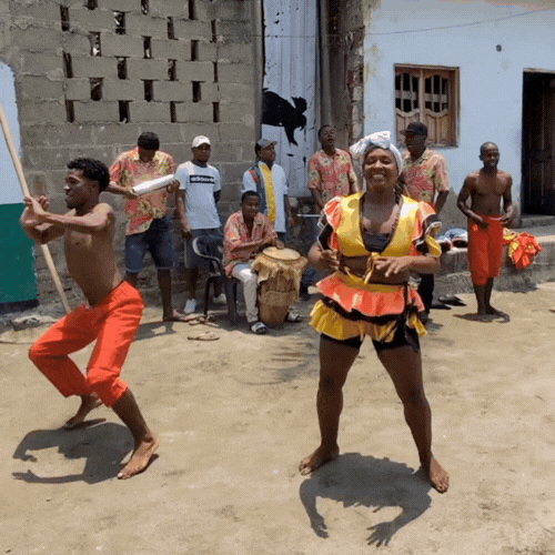 Group of people dancing and playing instruments on a dirt street in front of buildings.