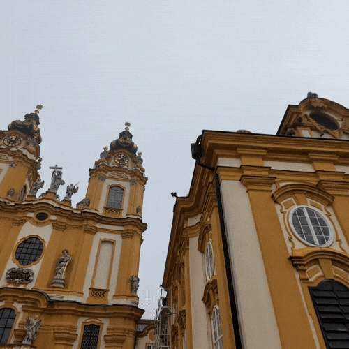View of Baroque-style yellow buildings with ornate architectural details and statues against a cloudy sky.