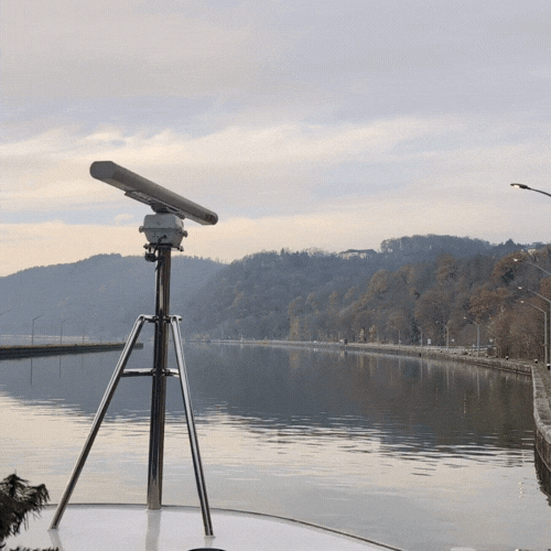 A telescope set up outdoors on a tripod by a river with hills and cloudy sky in the background.