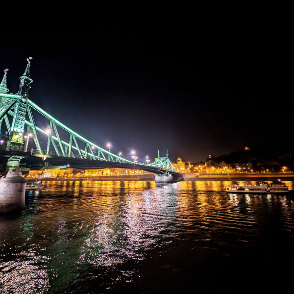 Night view of a illuminated bridge over a river, with reflections on the water and city lights in the background.