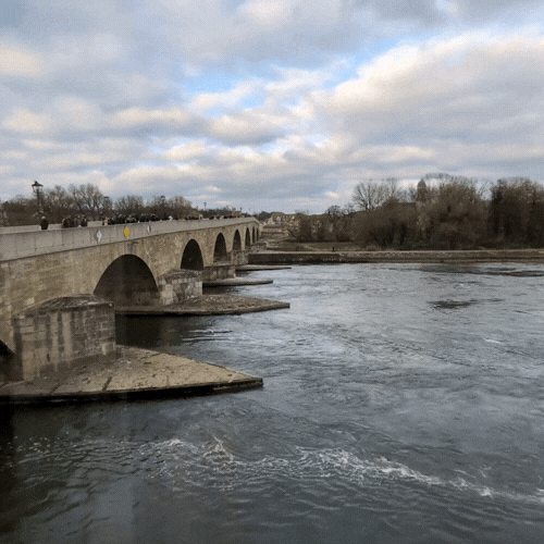 A stone bridge over a river with flowing water, trees in the background, and a cloudy sky above.
