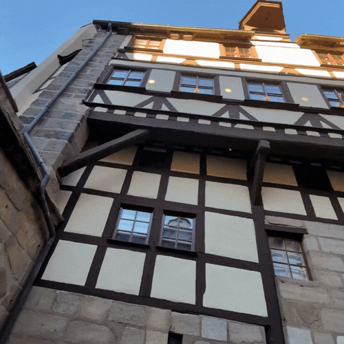 Close-up of a tall half-timbered building with exposed wooden beams and white plaster infill, showing windows and part of the stone foundation, under a clear blue sky.
