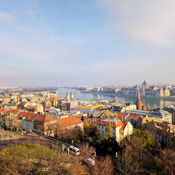 Panoramic view of a cityscape with a river flowing through the city, featuring historic buildings, churches with spires, and a mix of modern and old architecture under a partly cloudy sky.