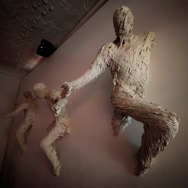 Large textured sculpture of a seated human figure with two smaller figures holding hands, all made from layered, white material, mounted on a wall.