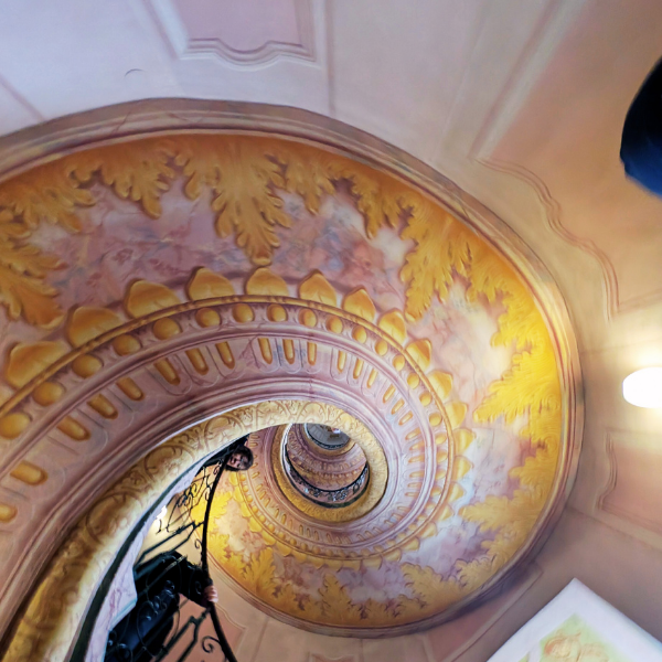 Photo of a spiral staircase viewed from the top, with ornate, colorful ceiling artwork featuring yellow leaves and decorative patterns.