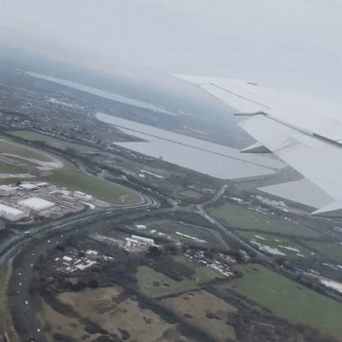 Aerial view of an airplane wing and a landscape with roads, buildings, and green fields below.