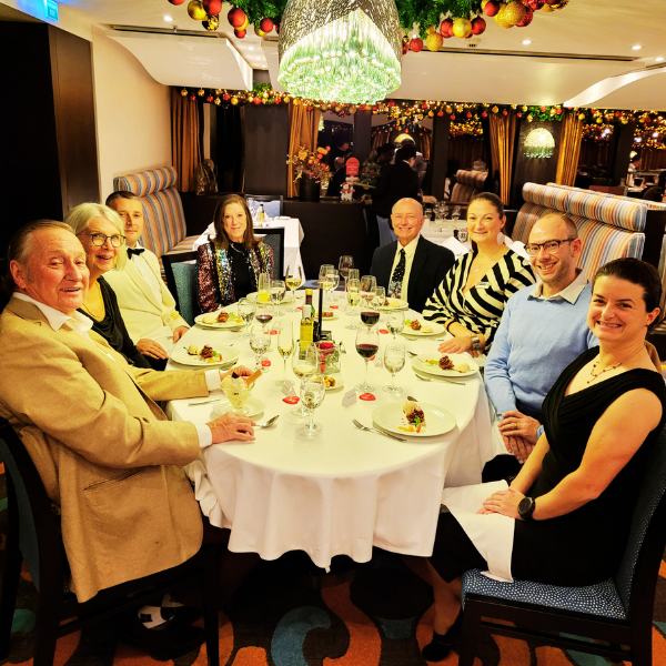 A group of seven people sitting around a round dinner table at a festive restaurant, celebrating with food and drinks, decorated with Christmas ornaments and greenery.