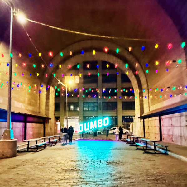 Night scene under a bridge with colorful string lights, benches, and a large illuminated "DUMBO" sign in the background.