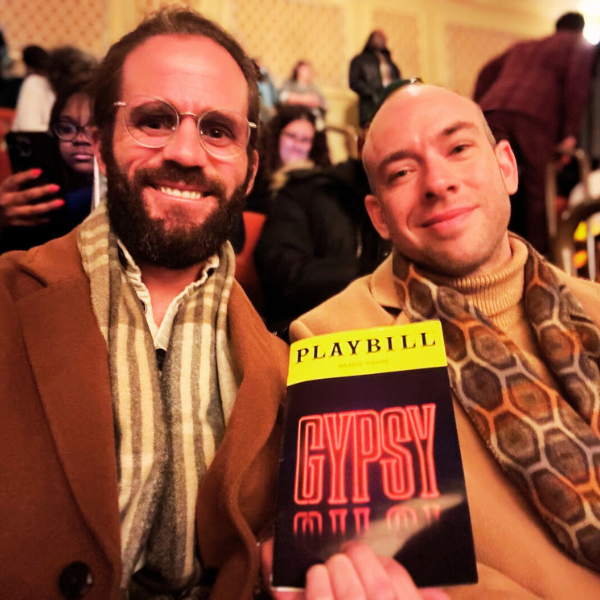 Two men sitting at a theater, smiling, holding a Playbill for the show 'Gypsy,' with audience members in the background.