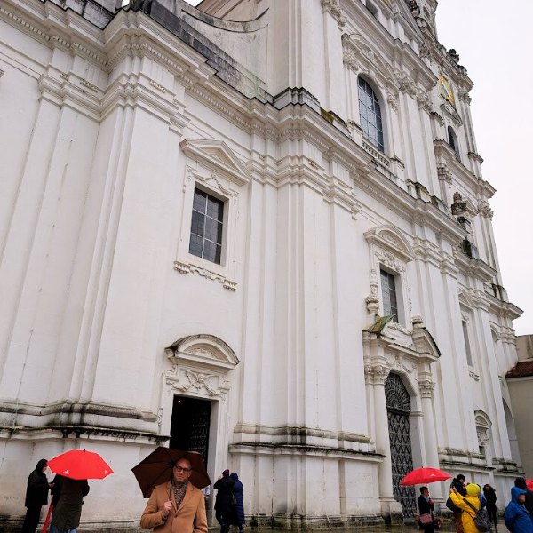 People with umbrellas walking outside a large, ornate white church or cathedral with baroque architectural details.