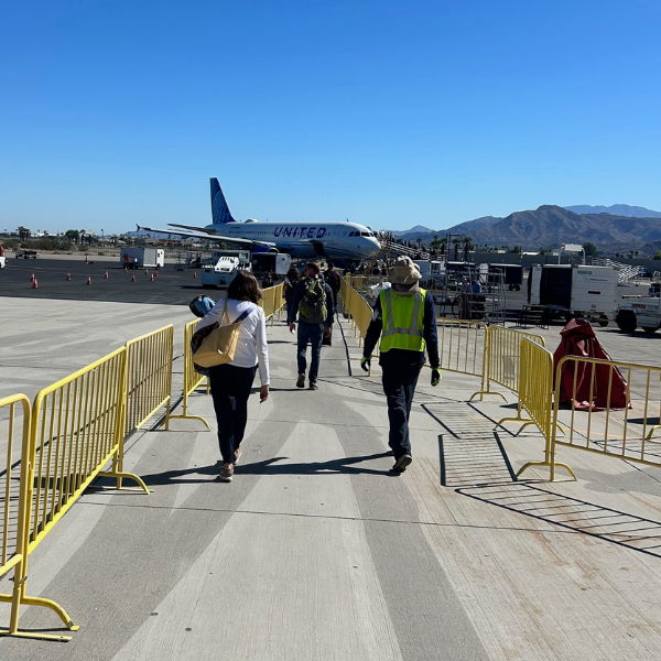 People walking on airport tarmac near a United Airlines airplane, with yellow barriers and mountains in the background.