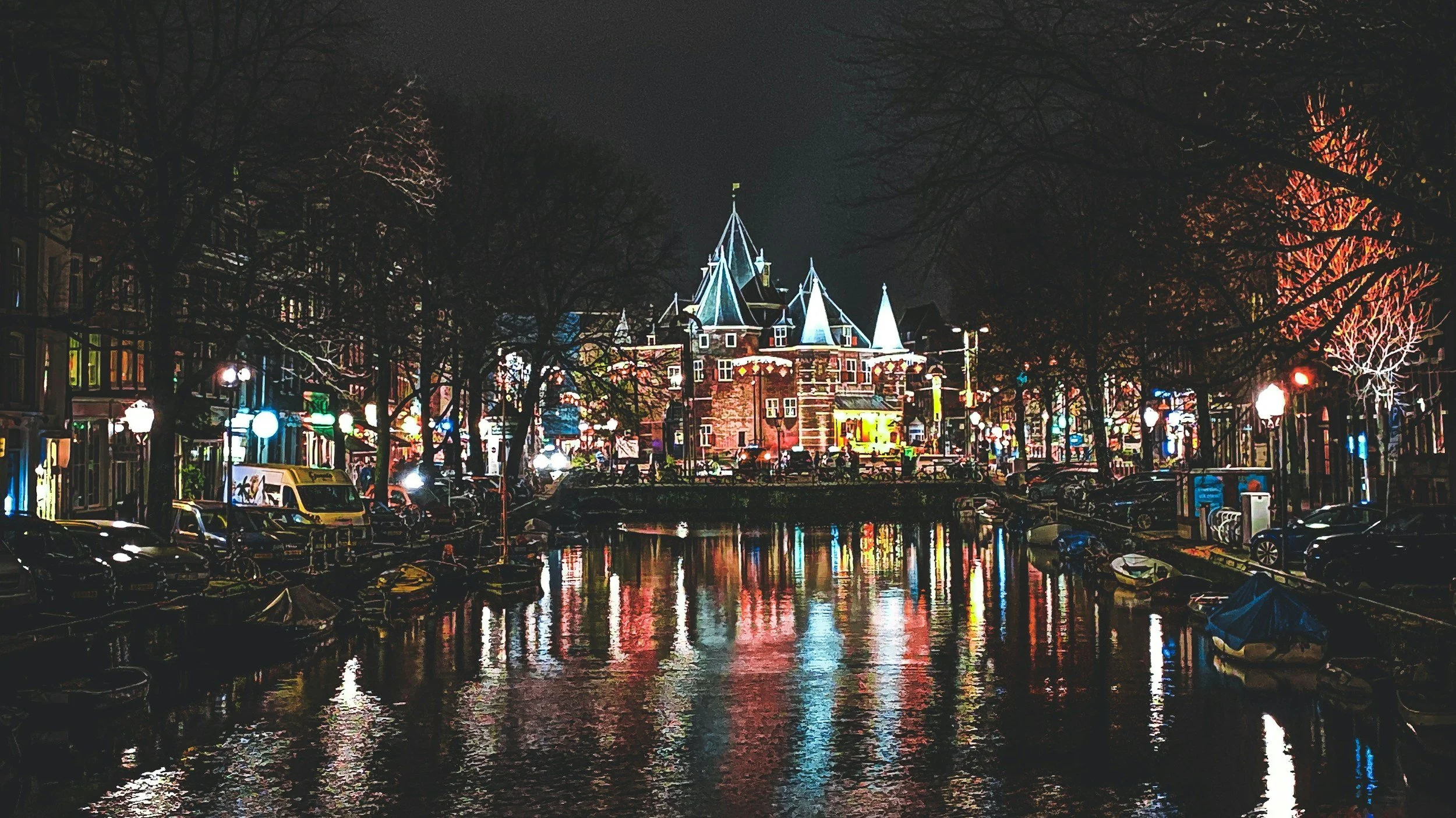 Night view of a canal with boats reflecting colorful city lights, surrounded by trees and historic buildings, including a castle-like structure illuminated in blue and yellow.