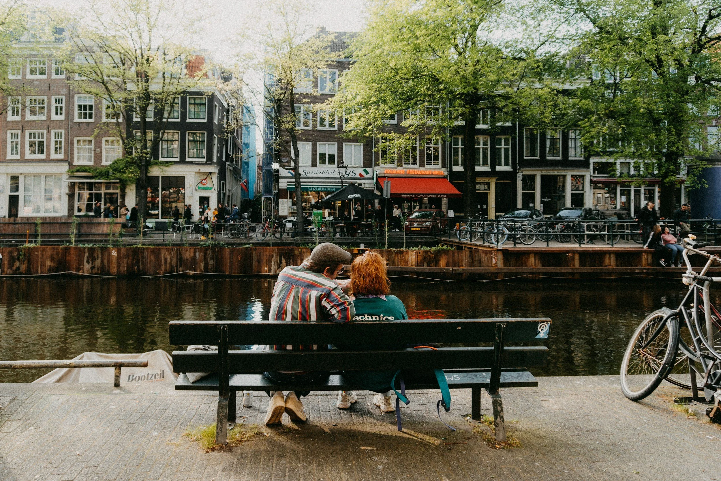 A view of a canal with two people sitting on a bench, boats, bicycles, and a row of buildings with shops and restaurants along the canal, and trees with fresh green leaves in a city.
