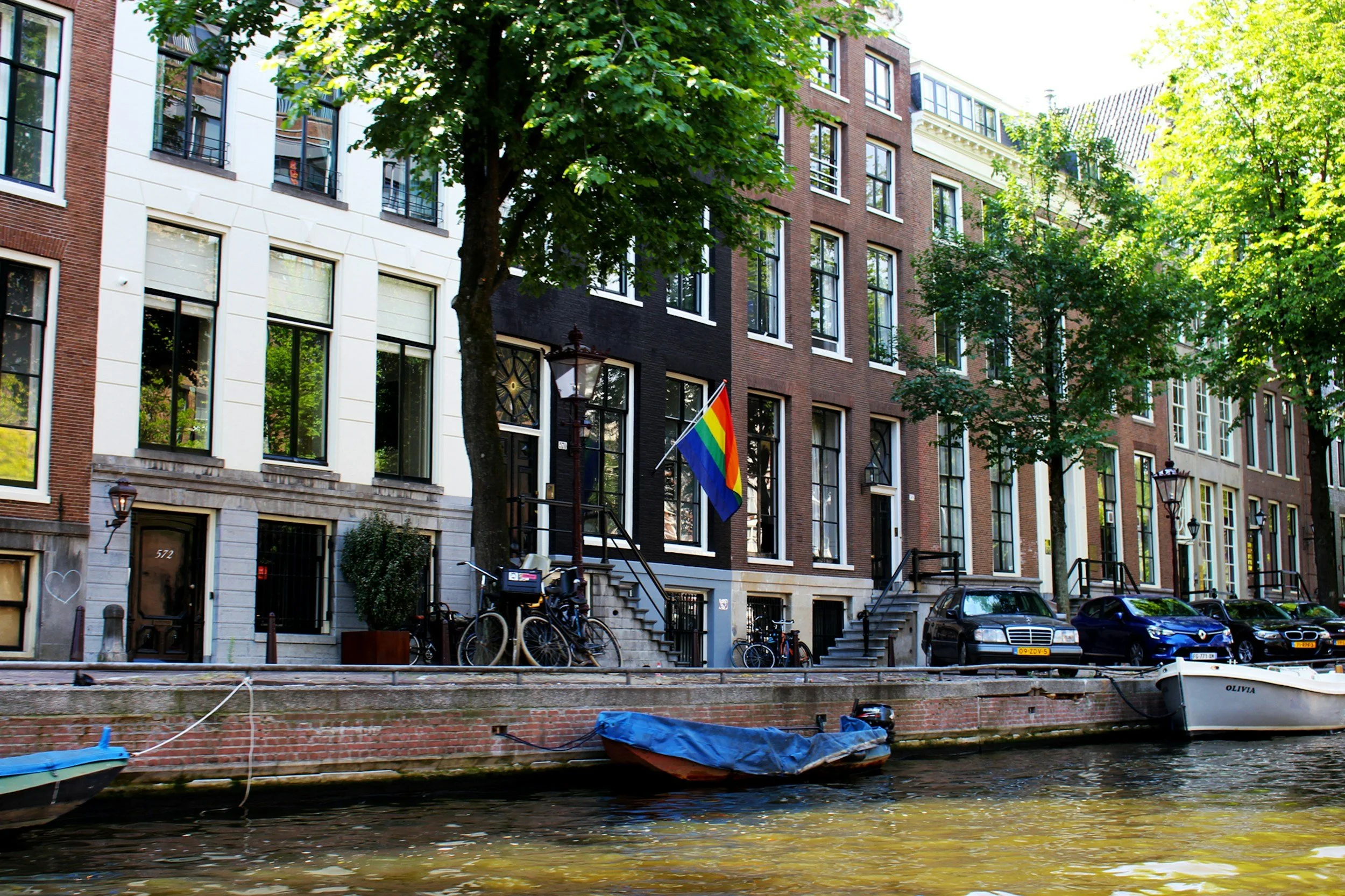 Amsterdam canal with boats docked along the water, row of narrow residential buildings with large windows and trees lining the street, rainbow pride flag hanging outside one building, bicycles parked on the sidewalk, parked cars along the street.