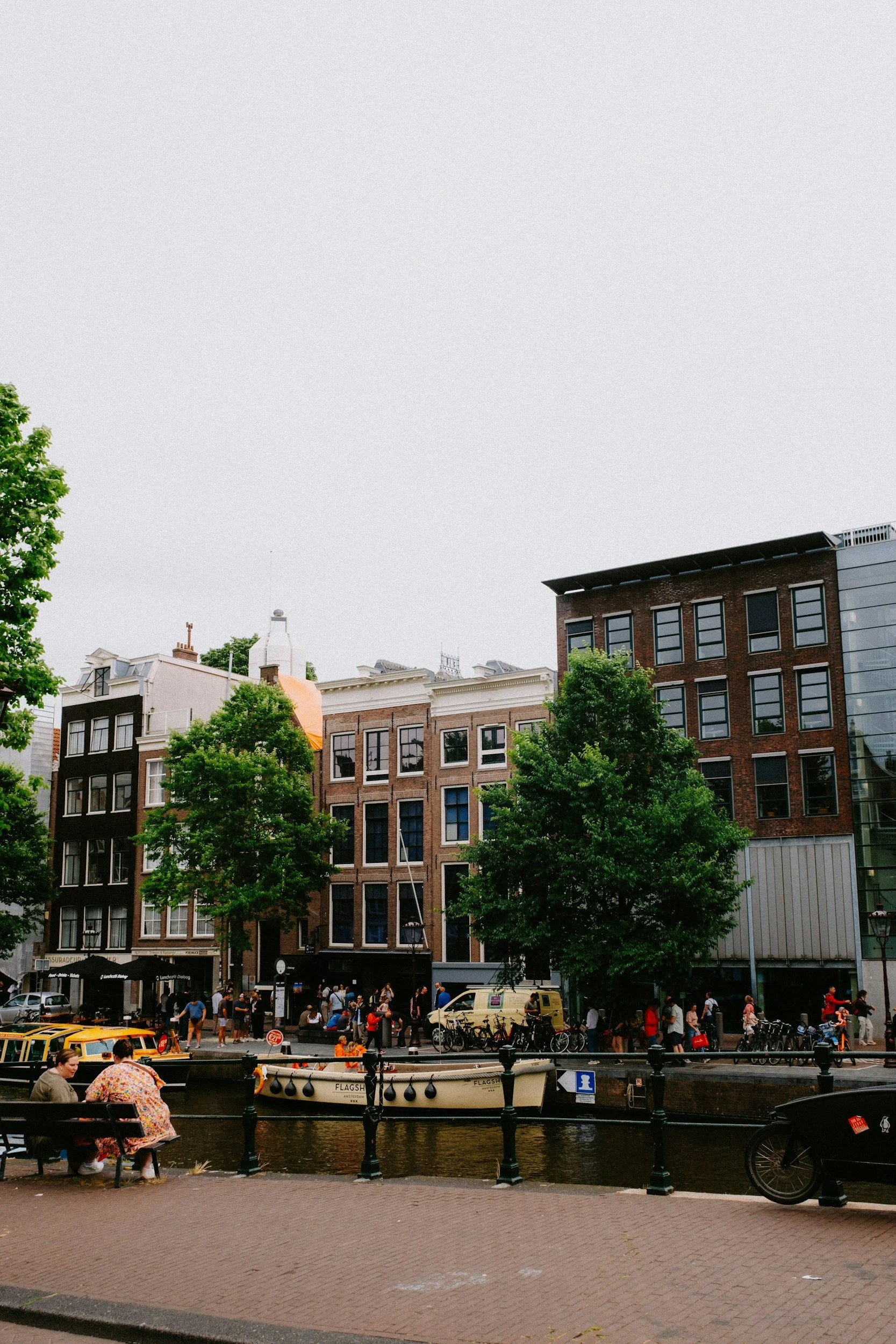 People walking along a canal in an urban cityscape with trees, traditional row houses, and modern buildings, with boats docked on the water and bicycles along the sidewalk.