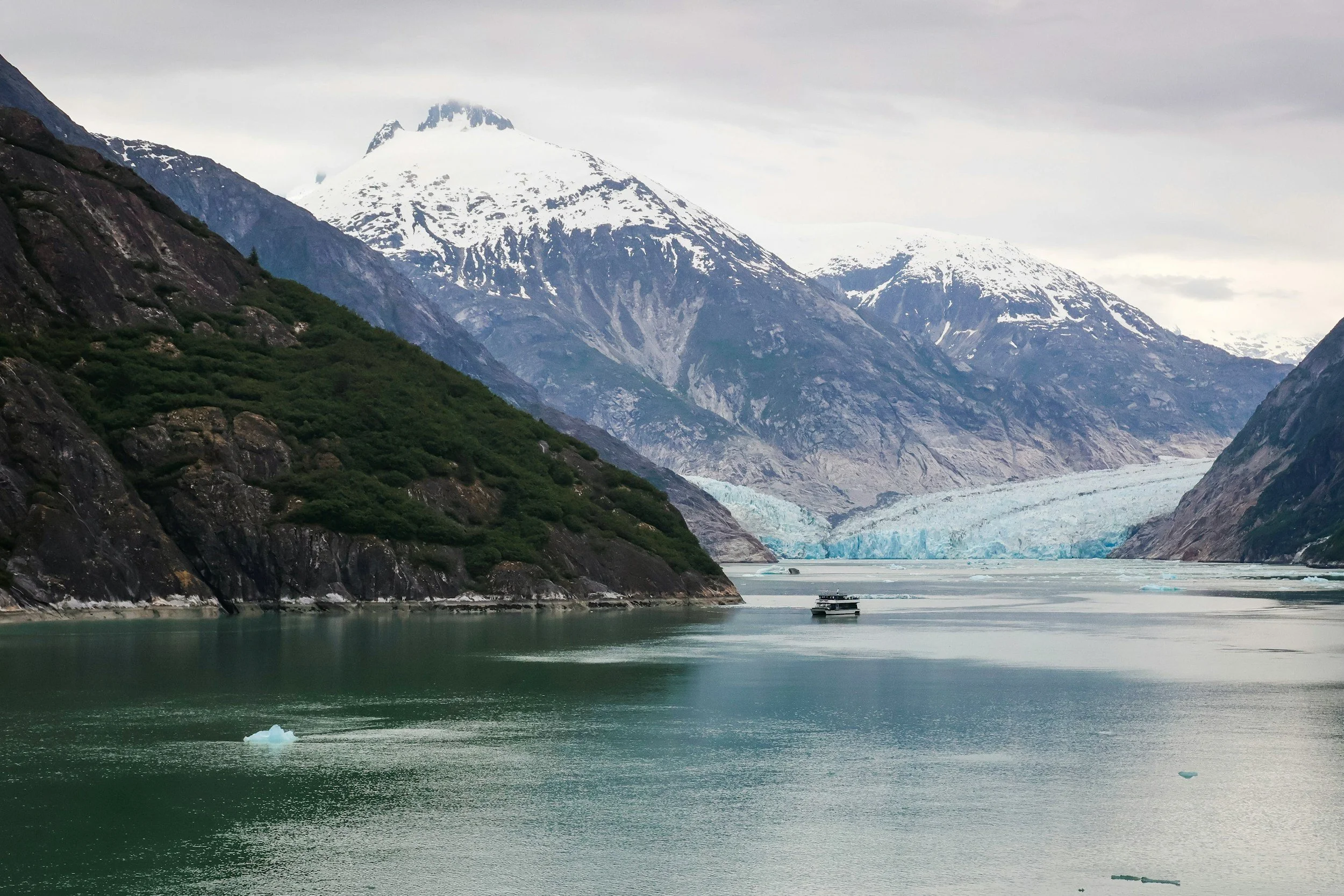 Mountains with snow and glaciers near a body of water with floating ice, small boat, and rocky, green slopes on the side.