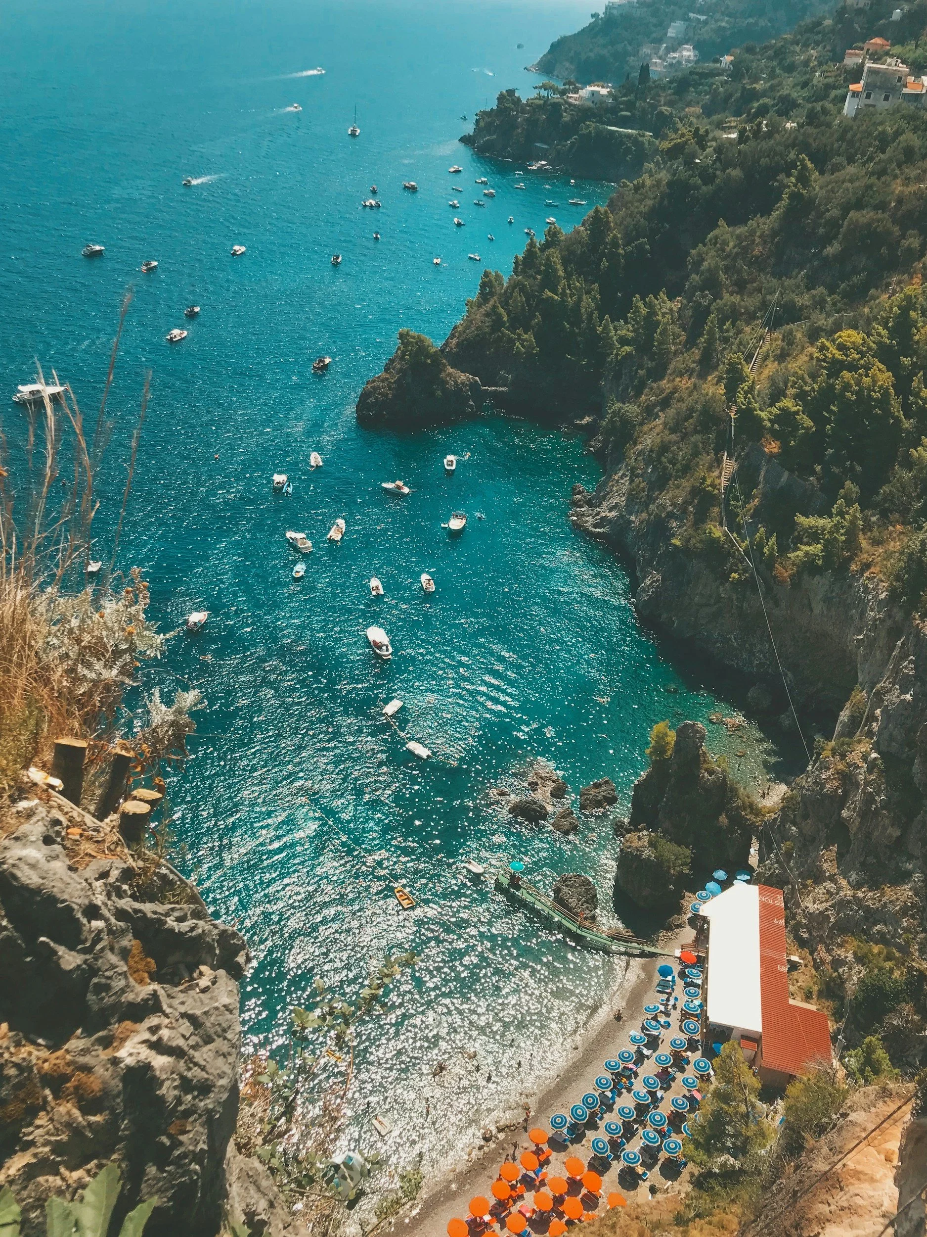 Beach with umbrellas and boats on the water, surrounded by cliffs and houses on the hillside.