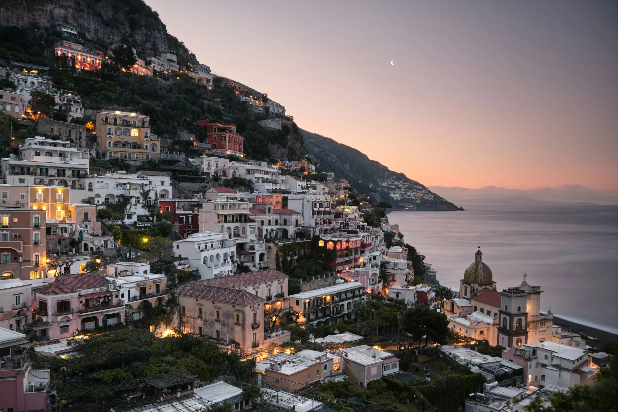 Houses and buildings on a hillside overlooking the ocean at sunset with a crescent moon in the sky.