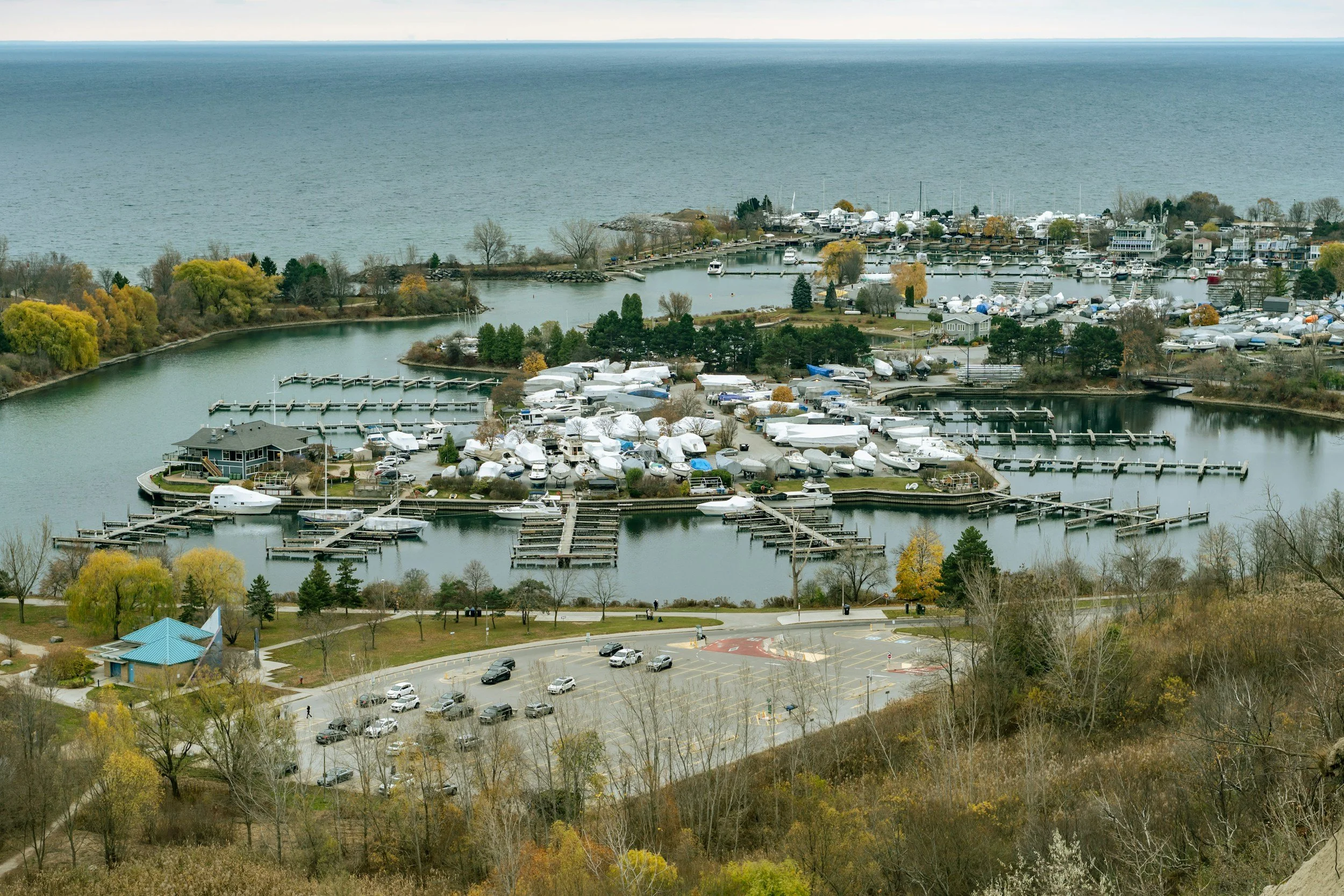 A harbor with boats docked, surrounded by trees with autumn foliage, and a distant large body of water.