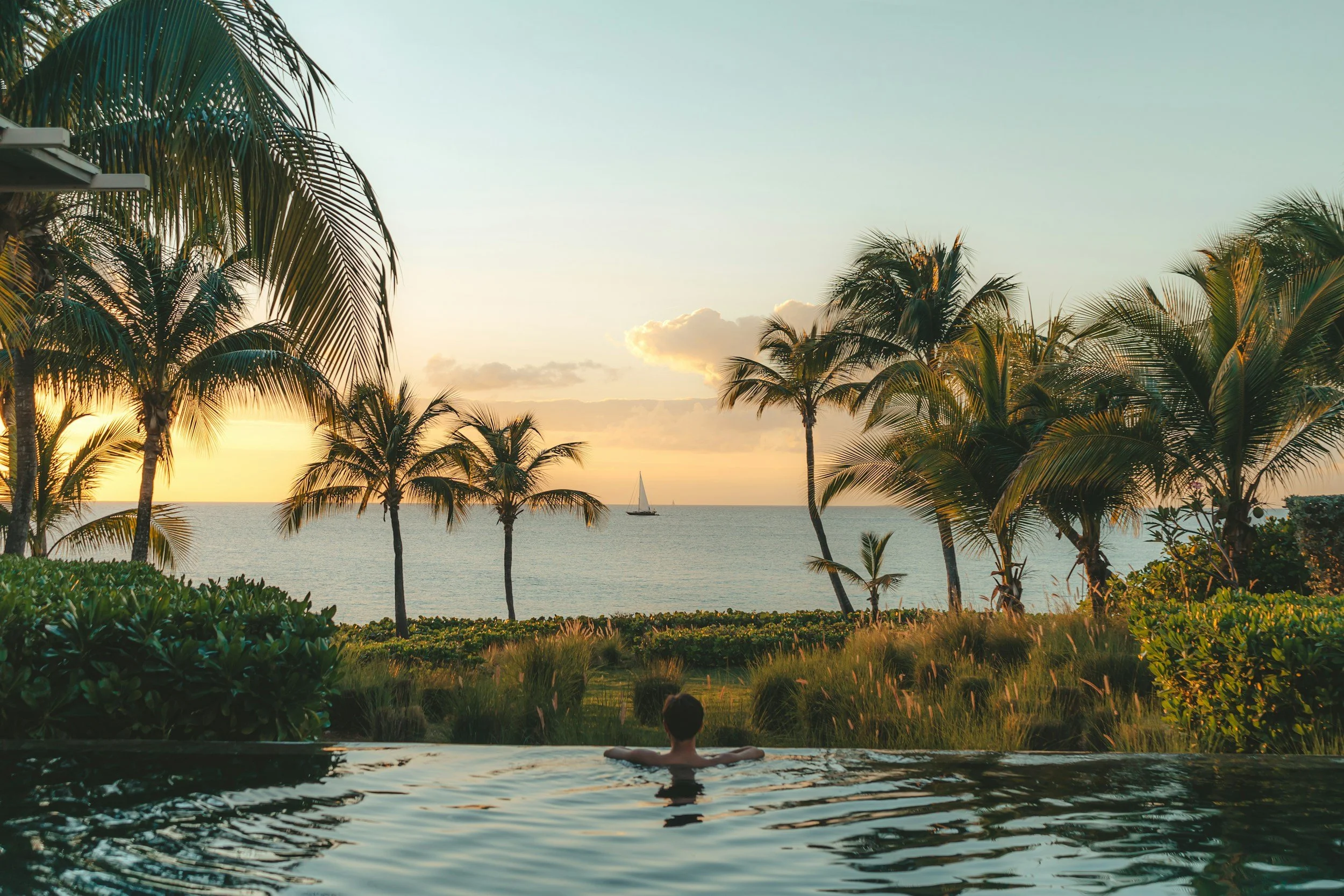 A person relaxing in a hot tub facing the ocean view with palm trees during sunset.