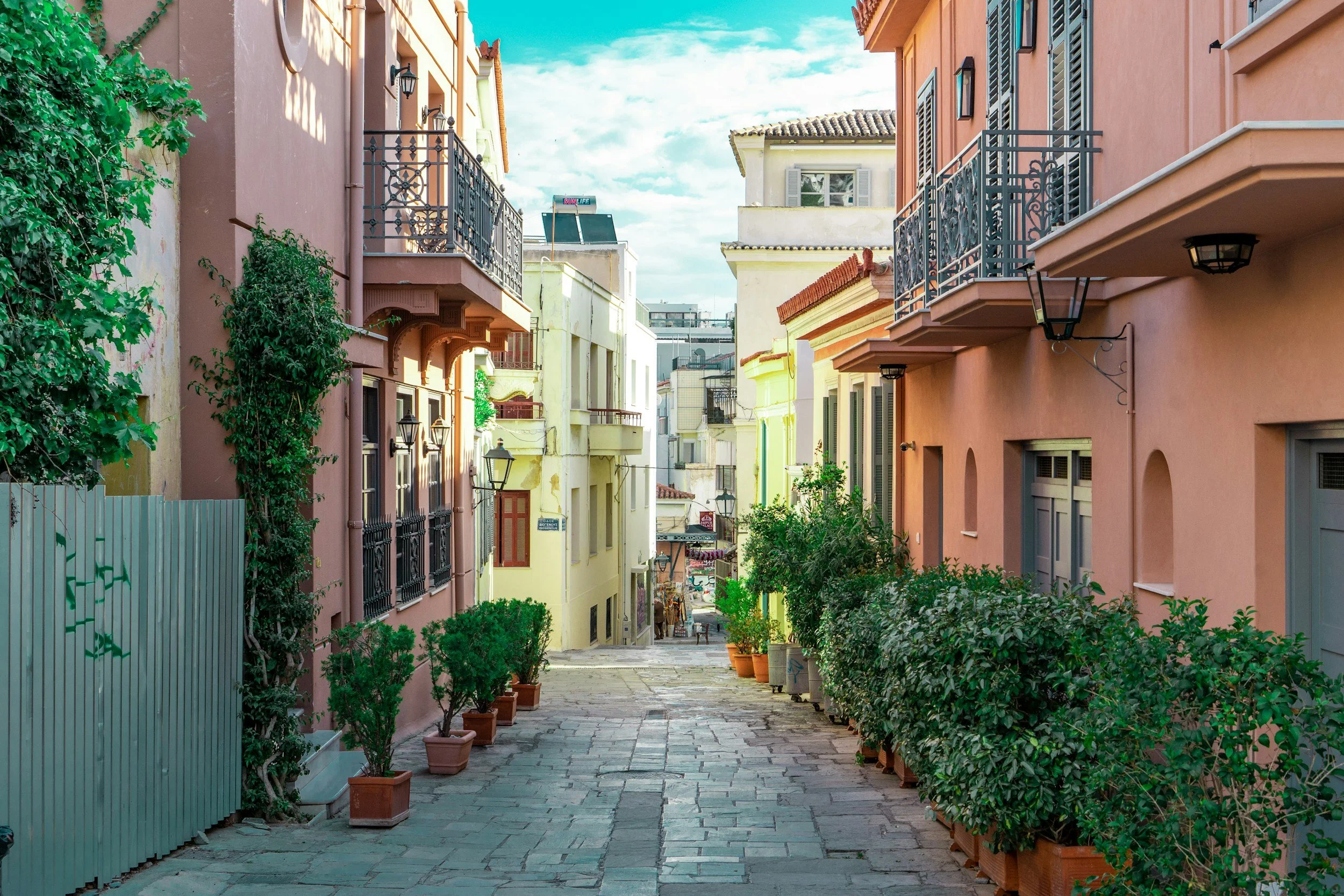 A colorful European street lined with pastel-colored buildings, potted plants, and trees, with a cobblestone pavement, under a partly cloudy blue sky.