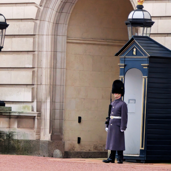 British guard in uniform standing outside a beige stone building with an archway, near a blue guardhouse and a lamp post.
