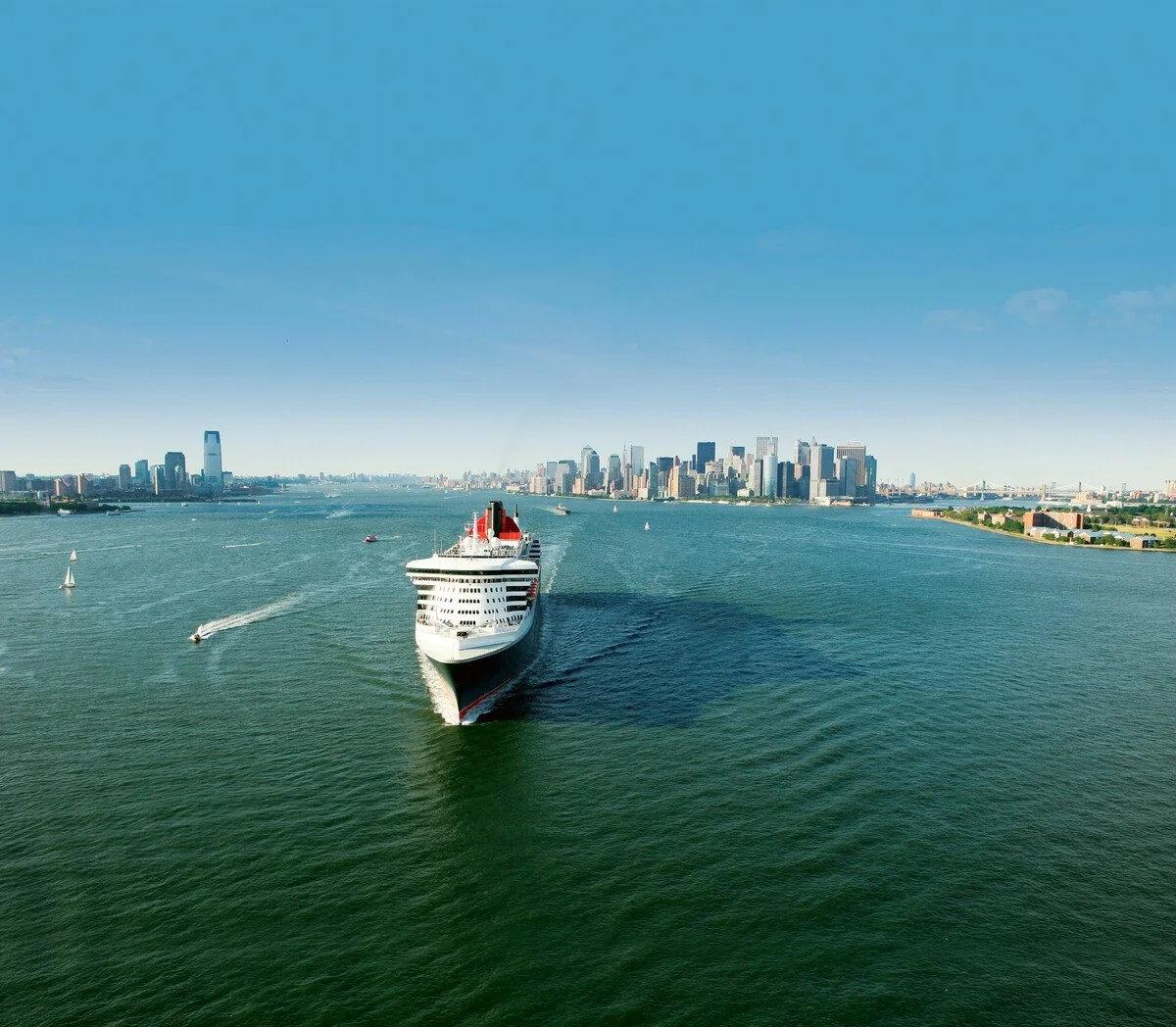 A large cruise ship sailing on a wide body of water with a city skyline in the background, featuring tall skyscrapers and a clear blue sky.