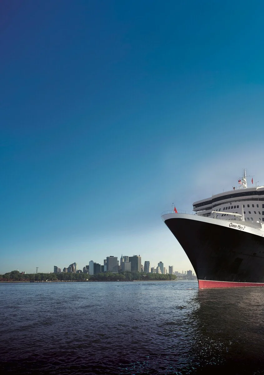 A large cruise ship named Queen Mary 2 docked on a body of water with a city skyline in the background.